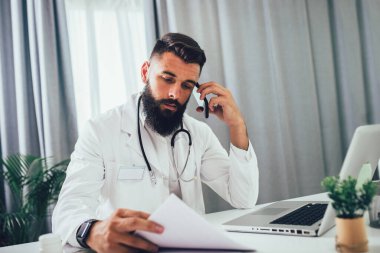Ofiste bilgisayarla çalışan ve telefon kullanan genç bir doktor..