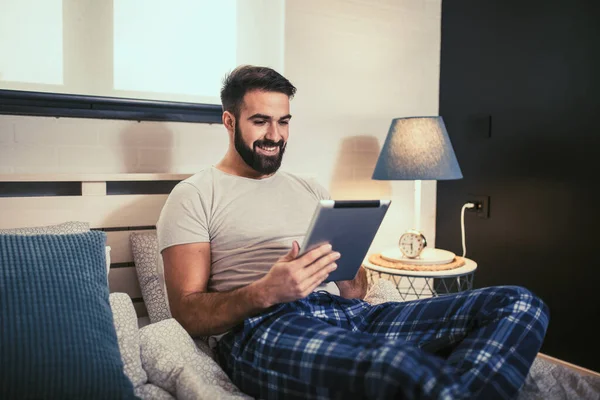 Handsome young man in pajamas using a digital tablet while lying in bed in the morning