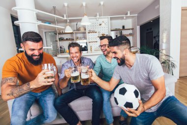 Erkekler televizyonda birlikte spor izleyip neşeli çığlıklar atıyorlar. Bir grup arkadaş kanepede oturup futbol maçı izliyorlar..