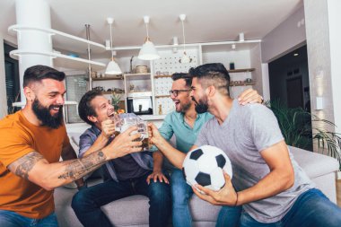 Erkekler televizyonda birlikte spor izleyip neşeli çığlıklar atıyorlar. Bir grup arkadaş kanepede oturup futbol maçı izliyorlar..