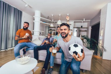 Erkekler televizyonda birlikte spor izleyip neşeli çığlıklar atıyorlar. Bir grup arkadaş kanepede oturup futbol maçı izliyorlar..