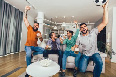 Erkekler televizyonda birlikte spor izleyip neşeli çığlıklar atıyorlar. Bir grup arkadaş kanepede oturup futbol maçı izliyorlar..