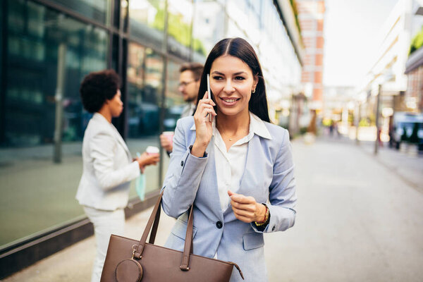 Portrait of beautiful smiling female in fashion office clothes talking on phone while standing outdoors.