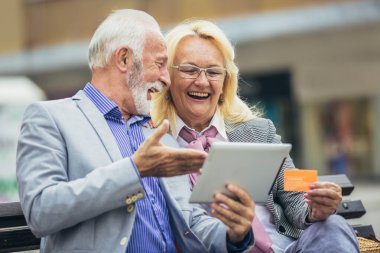 Yaşlı çift parkta bankta oturuyor ve tablet ve kredi kartıyla alışveriş yapıyor.