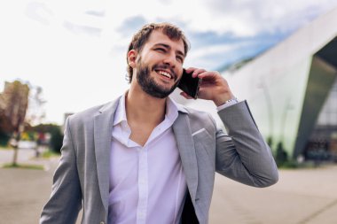 Zarif takım elbiseli kendine güvenen genç adam dışarıda duruyor, iş sırasında ya da seyahat sırasında akıllı telefondan konuşuyor..