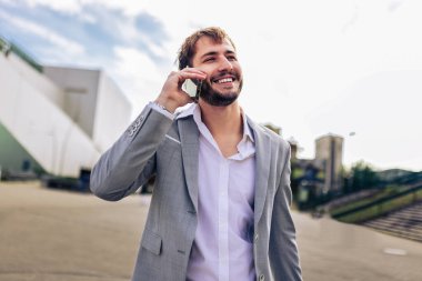 Zarif takım elbiseli kendine güvenen genç adam dışarıda duruyor, iş sırasında ya da seyahat sırasında akıllı telefondan konuşuyor..