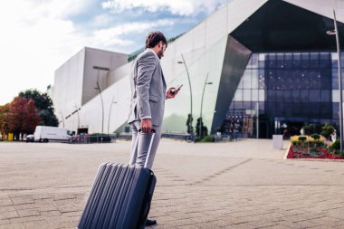 Havaalanı terminalinin önünde duran kendine güvenen iş adamı iş seyahatinde akıllı telefondan konuşuyor..