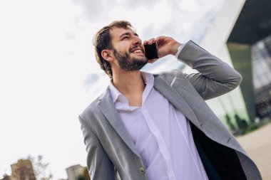 Zarif takım elbiseli kendine güvenen genç adam dışarıda duruyor, iş sırasında ya da seyahat sırasında akıllı telefondan konuşuyor..