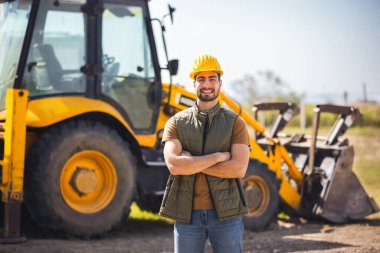 İnşaat alanında kazıcının önünde güvenle duran güvenlik donanımı giyen genç bir inşaat işçisi..