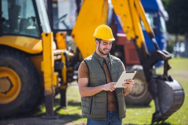 Kazıcının önünde elinde tabletle duran genç inşaat işçisi inşaat alanındaki planları veya verileri kontrol ediyor..