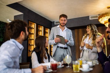 Kadın ve erkek iş yemeğinde meslektaşlarına bir tablet sunumu sunar. Takım dikkatle dinliyor, profesyonel odaklanmayı gevşek yemek atmosferiyle harmanlıyor..