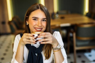 Kafedeki güzel genç bir kadın elinde kahve fincanı tutarken kameraya gülümsüyor, sıcaklığı, çekiciliği ve rahatlamış bir sabah havasını yakalıyor..