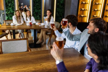 Üç adam bir barda bira içip masanın karşısındaki bir kadına neşeyle el sallıyor. Canlı atmosfer, sosyal etkileşim, eğlence ve gündelik arkadaşlık.