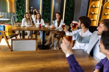 Üç adam bir barda bira içip masanın karşısındaki bir kadına neşeyle el sallıyor. Canlı atmosfer, sosyal etkileşim, eğlence ve gündelik arkadaşlık.