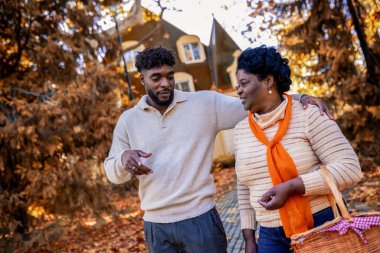 Afrika kökenli Amerikalı anne ve yetişkin oğul piknik çantasıyla sonbahar parkında yürüyorlar sıcak sonbahar gününün ve açık havada ailece eğleniyorlar..
