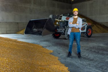 Güler yüzlü depo işçisi kollarını kavuşturup patinaj dümeni yükleyicisi önünde poz veriyor, güveni, sanayiyi ve tarımın sembolü.