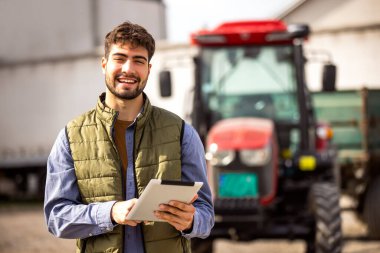 Traktörünün yanında duran ve tablete bakan genç traktör sürücüsü, modern tarım, teknoloji ve akıllı tarımın sembolü..