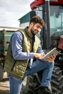Traktörünün yanında duran ve tablete bakan genç traktör sürücüsü, modern tarım, teknoloji ve akıllı tarımın sembolü..