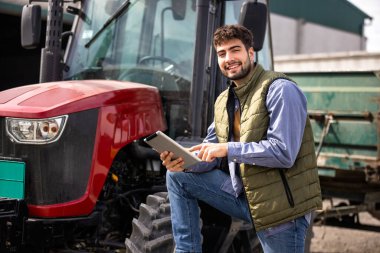 Traktörünün yanında duran ve tablete bakan genç traktör sürücüsü, modern tarım, teknoloji ve akıllı tarımın sembolü..