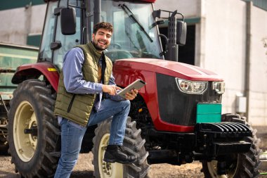 Traktörünün yanında duran ve tablete bakan genç traktör sürücüsü, modern tarım, teknoloji ve akıllı tarımın sembolü..