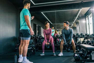 Spor salonundaki iki genç yetişkin ağırlıklı çömelme hareketleri yaparken, antrenör de zamanı ölçüyor, spor, güç ve eğitime odaklanıyor..