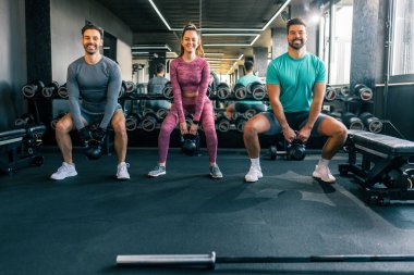 Spor salonunda iki genç adam ve bir kadın ağırlıklı çömelme hareketleri yapıyorlar. Güç, spor ve aktif antrenmanlara odaklanıyorlar..