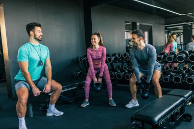 Spor salonundaki iki genç yetişkin ağırlıklı çömelme performansı sergilerken antrenör onlara rehberlik ediyor, fitness, güç ve düzgün antrenmanlara odaklanıyor..