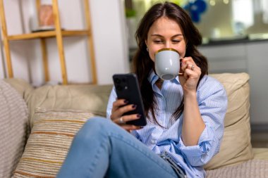 Rahat giyimli genç bir kadın, rahat bir oturma odasında huzurlu bir dinlenme zamanında rahat bir oturma odasında akıllı telefonunu kaydırırken kanepede rahatlar, kahve içer..