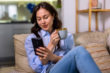 Rahat giyimli genç bir kadın, rahat bir oturma odasında huzurlu bir dinlenme zamanında rahat bir oturma odasında akıllı telefonunu kaydırırken kanepede rahatlar, kahve içer..