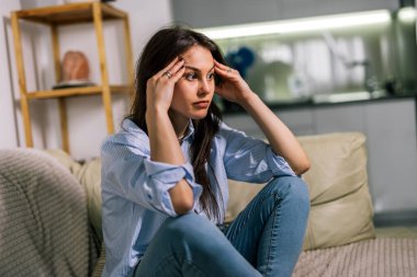 Genç bir kadın loş bir odada yalnız başına oturur, üzgün, gözyaşı dolu bir ifadeyle aşağıya bakar, depresyon ve duygusal mücadelenin sessiz bir anını yansıtır..