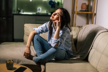 Genç bir kadın loş bir odada yalnız başına oturur, üzgün, gözyaşı dolu bir ifadeyle aşağıya bakar, depresyon ve duygusal mücadelenin sessiz bir anını yansıtır..