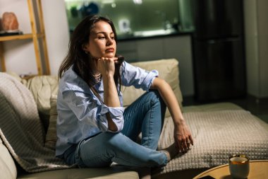 Genç bir kadın loş bir odada yalnız başına oturur, üzgün, gözyaşı dolu bir ifadeyle aşağıya bakar, depresyon ve duygusal mücadelenin sessiz bir anını yansıtır..