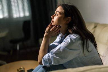 Genç bir kadın loş bir odada yalnız başına oturur, üzgün, gözyaşı dolu bir ifadeyle aşağıya bakar, depresyon ve duygusal mücadelenin sessiz bir anını yansıtır..