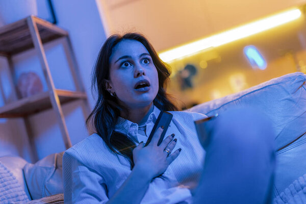 Молодая женщина сидит в своей гостиной, освещенной синим светом от телевизора, явно напуганная и напряженная, пока смотрит фильм ужасов.