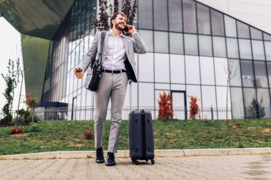 Dışarıda telefonla konuşan, bavul ve kahve fincanı taşıyan meşgul bir işadamı. Kentsel bir ortamda seyahat ederken ya da seyahat ederken modern profesyonel çoklu görev.