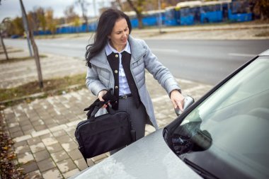 Orta yaşlı bir kadın arabasındaki çizikleri inceliyor, hasarı dikkatlice kontrol ediyor ve durumu değerlendiriyor..