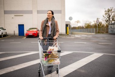 Kırklı yaşlarda bir kadın süpermarketin önüne alışveriş arabası itiyor, mağazaya girmeden önce otoparkta geziniyor..