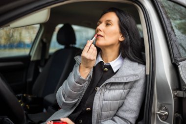 Orta yaşlı bir kadın ruj tutuyor ve araba kullanmaya hazırlanırken güneşlik aynasında yansımasını kontrol ediyor..