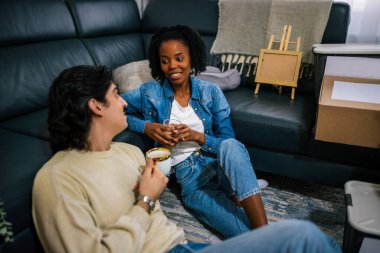 Karışık çift yeni dairelerinin oturma odasında mutlu bir şekilde oturuyorlar, etrafı taşınan kutular ve gitarlarla çevrili, bir fincan kahve tutan adam, ilk evlerini kutluyor..