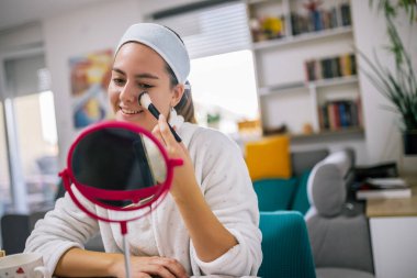 Bornozlu genç bir kadın evde oturuyor, küçük bir aynaya bakarken makyaj yapıyor..