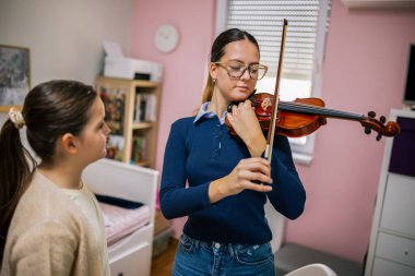 Eğitmen, evde müzik dersi sırasında kıza kemanı nasıl tutacağını ve yayı doğru şekilde hareket ettireceğini gösterir..