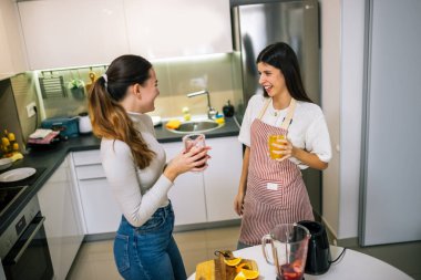İki genç kadın modern bir mutfakta taze meyve suyu ve meyve sularının tadını çıkarıyorlar. Gülümsüyorlar ve sağlıklı, renkli bir içki anı paylaşıyorlar..