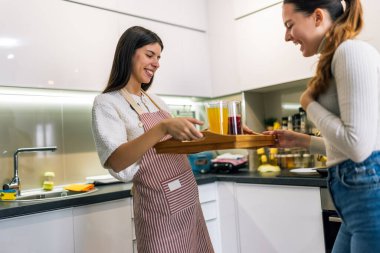 Önlüklü genç bir kadın, modern bir mutfaktaki arkadaşına taze meyve suyu ve kurabiyelerle dolu bir tabak getiriyor. Neşeli ve sağlıklı bir atıştırma anını paylaşıyor..