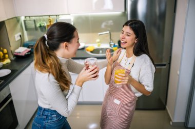 İki genç kadın modern bir mutfakta taze meyve suyu ve meyve sularının tadını çıkarıyorlar. Gülümsüyorlar ve sağlıklı, renkli bir içki anı paylaşıyorlar..