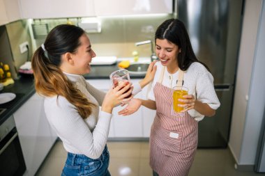 İki genç kadın modern bir mutfakta taze meyve suyu ve meyve sularının tadını çıkarıyorlar. Gülümsüyorlar ve sağlıklı, renkli bir içki anı paylaşıyorlar..