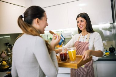 Önlüklü genç bir kadın, modern bir mutfaktaki arkadaşına taze meyve suyu ve kurabiyelerle dolu bir tabak getiriyor. Neşeli ve sağlıklı bir atıştırma anını paylaşıyor..