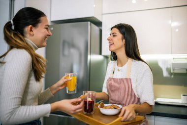 Önlüklü genç bir kadın, modern bir mutfaktaki arkadaşına taze meyve suyu ve kurabiyelerle dolu bir tabak getiriyor. Neşeli ve sağlıklı bir atıştırma anını paylaşıyor..