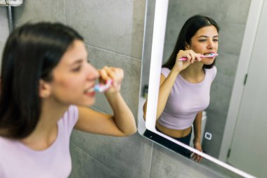 Genç bir kadın dişlerini banyoda büyük bir aynanın önünde fırçalıyor, ağız sağlığını ve sağlıklı gülümsemesini koruyor..