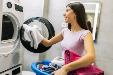 Genç bir kadın banyodaki çamaşır makinesinden temiz havluları mutlulukla alıyor, yeni yıkanmış çamaşırların tadını çıkarıyor..