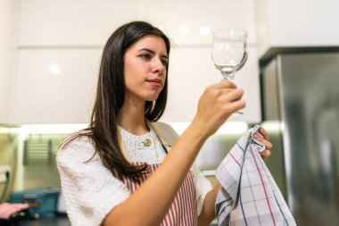 Önlüklü genç bir kadın mutfağında bir bezle şarap bardağını temizliyor. Ev işlerini yaparken temizlik yapıyor..